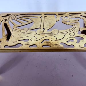 May include: A gold-coloured metal trivet with a rectangular shape and intricate cut-out design featuring a ship, waves, and sun. The trivet has four claw-footed legs and is likely used to protect surfaces from hot items.