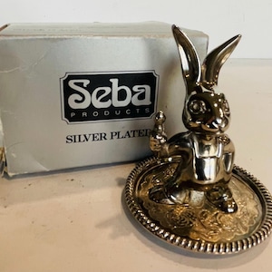 Könnte beinhalten: Versilberte Hasenfigur auf einer dekorativen Platte mit Perlenrand. Daneben eine Schachtel mit dem Text "Seba Products Silver Plated". Der Hase hält einen kleinen Gegenstand.