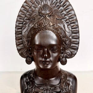 May include: A dark brown wooden bust of a woman with closed eyes, wearing a detailed headdress and earrings. The bust features intricate carvings on the headpiece, earrings, and clothing, showcasing a traditional design.