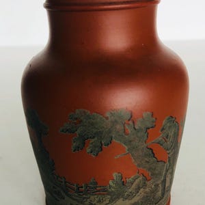 Könnte beinhalten: Eine terrakottafarbene Vase mit detailliertem Landschaftsdesign. Die Vase hat einen breiten Sockel, einen zylindrischen Körper und einen leicht ausgestellten Rand. Das Landschaftsdesign ist in Grautönen gehalten und zeigt Bäume, Figuren und einen Zaun.