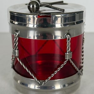 May include: A silver and red glass jar shaped like a drum with a silver lid and two drumsticks. The jar has a red glass body with silver bands around it.