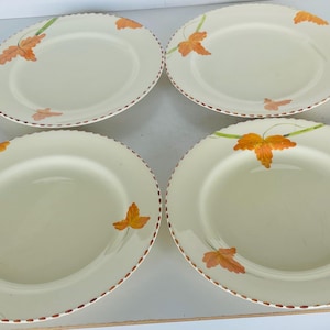 May include: Four off-white ceramic plates with a scalloped edge and a brown trim. Each plate features a design of orange and yellow leaves with green stems. The plates are arranged on a white surface.