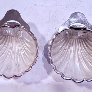 May include: Two shell-shaped dishes with silver-coloured metal trim. The dishes have a scalloped edge and a fan-like pattern inside. They are likely for serving small portions of food or as decorative items.