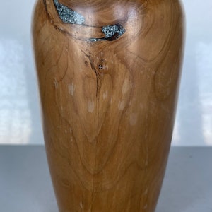 Könnte beinhalten: Eine Holzvase mit hellbrauner Oberfläche und einem einzigartigen Einlegemuster aus Silber und Grau. Die Vase hat eine glatte, abgerundete Form und eine leicht ausgestellte Öffnung.