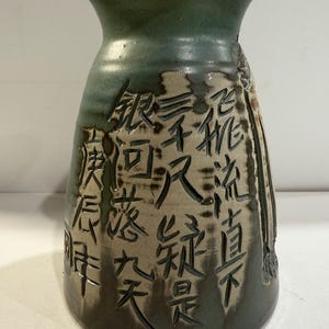 May include: A green ceramic vase with a wide mouth and a narrow base. The vase is decorated with carved Chinese characters.