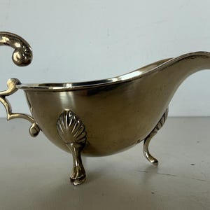May include: Silver gravy boat with a curved spout and ornate handle. The boat has three small feet and a decorative shell design on the side. A classic serving piece for sauces and gravies.