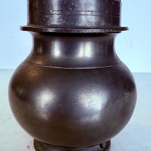 Könnte beinhalten: Ein dunkler, metallisch gefärbter Topf mit Deckel. Der Topf hat einen abgerundeten Körper, einen ausgestellten Hals und einen breiten Boden. Der Deckel sitzt oben auf dem Topf und hat einen Griff. Der Topf scheint aus Metall zu sein und hat eine leicht strukturierte Oberfläche.