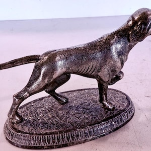 May include: A silver-toned dog statue in a poised stance, with one front leg raised. The dog is on an oval base with a textured pattern and decorative border. The sculpture has a detailed, realistic appearance.