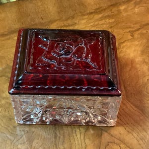 May include: A rectangular, ruby-red glass jewelry box with a clear, faceted base. The lid features a raised rose design. The box is likely vintage and sits on a wooden surface.