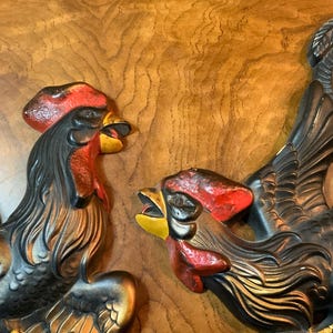 May include: Three-dimensional rooster wall art with a rustic aesthetic. The roosters feature black bodies, red combs, and yellow beaks. The background is a warm, wood-grain surface, enhancing the vintage appeal of the decorative pieces.