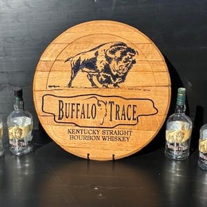 以下が含まれることがあります： 木製の樽の頭にバイソンの彫刻と「BUFFALO TRACE KENTUCKY STRAIGHT BOURBON WHISKEY」の文字が刻まれています。ラベルの付いた4本の空のガラス製ウイスキーボトルが樽の頭の前にあります。