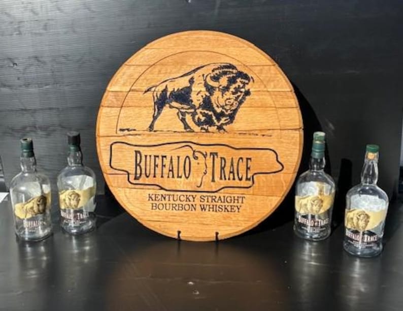 以下が含まれることがあります： 木製の樽の頭にバイソンの彫刻と「BUFFALO TRACE KENTUCKY STRAIGHT BOURBON WHISKEY」の文字が刻まれています。ラベルの付いた4本の空のガラス製ウイスキーボトルが樽の頭の前にあります。