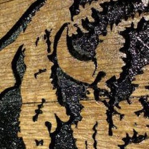 以下が含まれることがあります： 木製の表面のクローズアップ。彫刻されたデザインが施されています。黒と金のアクセントで、トラの顔の精巧な画像が描かれています。木目が見え、アートワークに質感を加えています。