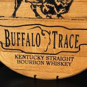 以下が含まれることがあります： 「BUFFALO TRACE」の文字がスタイリッシュなフォントで書かれた木製樽の一部をクローズアップ。下には「KENTUCKY STRAIGHT BOURBON WHISKEY」と書かれています。テキストの上にはバッファローの画像がエッチングされています。木材は自然なライトブラウン色です。