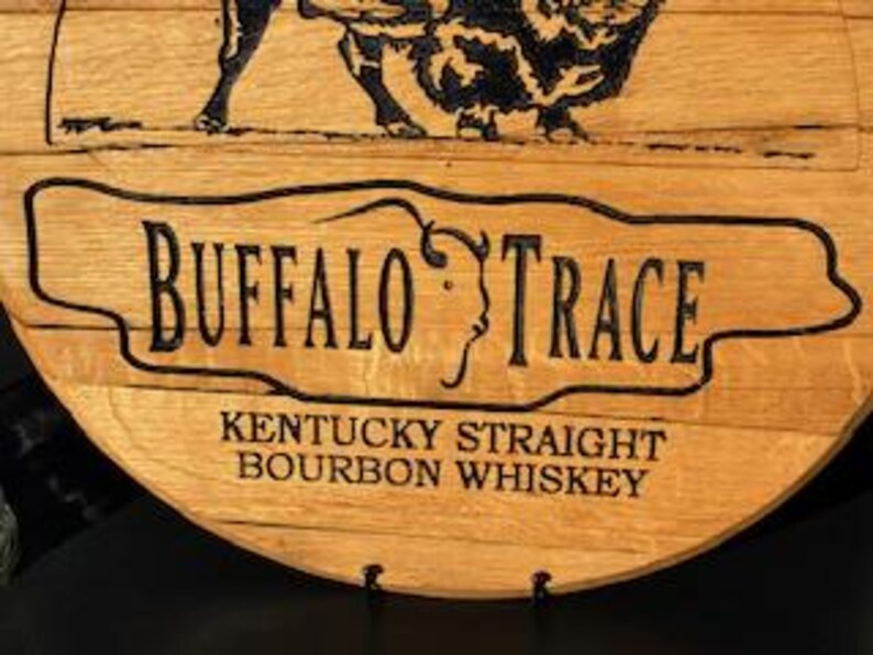以下が含まれることがあります： 「BUFFALO TRACE」の文字がスタイリッシュなフォントで書かれた木製樽の一部をクローズアップ。下には「KENTUCKY STRAIGHT BOURBON WHISKEY」と書かれています。テキストの上にはバッファローの画像がエッチングされています。木材は自然なライトブラウン色です。