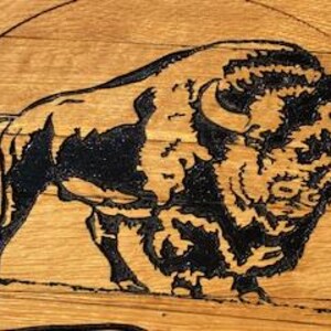 以下が含まれることがあります： 明るい茶色の木目模様の背景に、黒いバイソンの詳細な木彫り。 バイソンは横顔で描かれ、角、粗い毛皮、力強い姿勢を示しています。 彫刻は複雑で芸術的です。