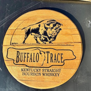 以下が含まれることがあります： 「Buffalo Trace」の文字とバイソンのイラストが描かれた丸い木製の看板。看板には「Kentucky Straight Bourbon Whiskey」の文字も含まれています。看板は明るい茶色の木材で作られています。