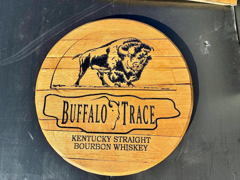 以下が含まれることがあります： 「Buffalo Trace」の文字とバイソンのイラストが描かれた丸い木製の看板。看板には「Kentucky Straight Bourbon Whiskey」の文字も含まれています。看板は明るい茶色の木材で作られています。