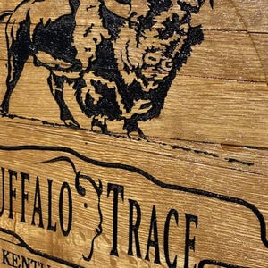 以下が含まれることがあります： 黒いバッファローのイラストと、曲がったバナーに「BUFFALO TRACE」という文字が描かれた木製の看板。看板には、ケンタッキー・ストレート・バーボン・ウイスキーに関するテキストも含まれています。