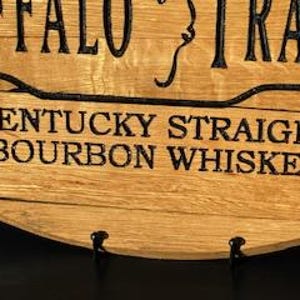 以下が含まれることがあります： 上部に「BUFFALO TRACE」の文字が入った木製の看板のクローズアップ。下には「KENTUCKY STRAIGHT BOURBON WHISKEY」と太字の黒いフォントで書かれています。看板は木目がはっきり見える明るい茶色の木材で作られています。