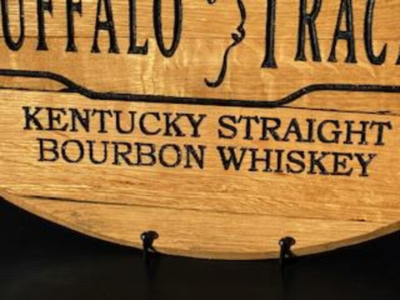 以下が含まれることがあります： 上部に「BUFFALO TRACE」の文字が入った木製の看板のクローズアップ。下には「KENTUCKY STRAIGHT BOURBON WHISKEY」と太字の黒いフォントで書かれています。看板は木目がはっきり見える明るい茶色の木材で作られています。