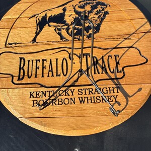 以下が含まれることがあります： 「BUFFALO TRACE」と「KENTUCKY STRAIGHT BOURBON WHISKEY」の文字が刻まれた丸い木製の看板。テキストの上には黒いバイソンのシルエットがあります。看板の前には金属製のスタンドがあります。