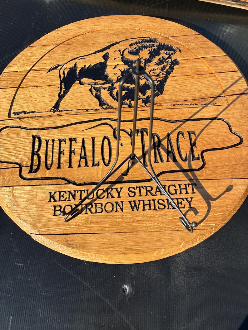 以下が含まれることがあります： 「BUFFALO TRACE」と「KENTUCKY STRAIGHT BOURBON WHISKEY」の文字が刻まれた丸い木製の看板。テキストの上には黒いバイソンのシルエットがあります。看板の前には金属製のスタンドがあります。