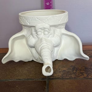 Puede incluir: Maceta de cerámica blanca con forma de cabeza de elefante con orejas grandes. La maceta tiene una superficie texturizada y una abertura ancha en la parte superior.