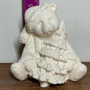 Könnte beinhalten: Unbemalte Keramik-Bärenfigur, die einen Hut trägt und einen Weihnachtsbaum hält. Der Bär ist etwa 13 cm groß. Die Figur ist zum Bemalen und Dekorieren bestimmt.