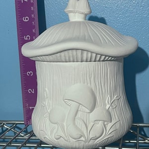 Puede incluir: Jarrón de cerámica blanca en forma de hongo con tapa. El jarrón tiene una superficie texturizada y presenta un diseño de hongos y hojas.