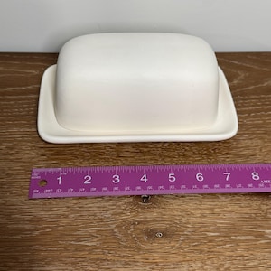 May include: A white ceramic butter dish with a lid, measuring approximately 6 inches long. The dish is rectangular with rounded edges and sits on a matching base. The butter dish is on a wooden surface.