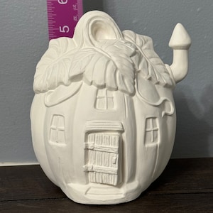 May include: White ceramic pumpkin house with a door, windows, and a chimney. The house is decorated with leaves and has a whimsical design.