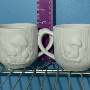 Könnte beinhalten: Zwei weiße Keramikbecher mit einem Pilz-Design. Die Becher haben eine strukturierte Oberfläche, die an Holz erinnert. Die Becher sind durch einen einzigen Griff verbunden.