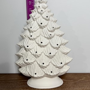 May include: A white ceramic Christmas tree with a textured surface and small black holes for lights. The tree is approximately 12 inches tall and sits on a round base. A ruler is placed next to the tree.