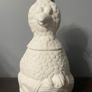 May include: A white ceramic bird-shaped cookie jar with a lid. The jar has a detailed, textured surface and a whimsical design. The bird's head is upright, with large eyes and a beak. The jar is approximately 13 inches tall.