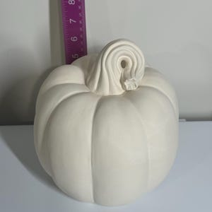 May include: A cream-colored ceramic pumpkin with a detailed stem. The pumpkin is approximately 6 inches tall. The pumpkin has vertical ridges and a curled stem, perfect for fall decor or crafting projects.