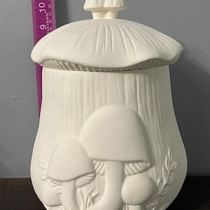 Puede incluir: Un tarro blanco de cerámica con forma de hongo y tapa. El tarro presenta diseños de hongos en relieve en la parte delantera y una superficie texturizada. La tapa también tiene forma de hongo. El tarro mide aproximadamente 28 cm de alto.