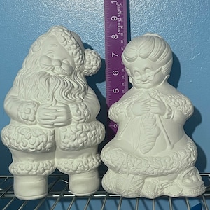 May include: Two white ceramic figurines, one of Santa Claus and one of a woman knitting. Both figurines are standing on a metal shelf.