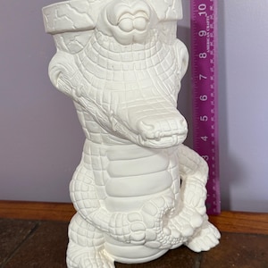 May include: White ceramic alligator planter with a textured surface and a wide opening at the top. The alligator is sitting with its legs crossed and its tail curled around its body.