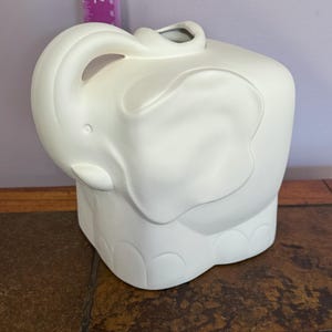 Op de afbeelding: Witte keramische olifantvormige plantenbak met een vierkante basis. De slurf van de olifant is opgerold en de plantenbak heeft een gat aan de bovenkant om te planten.