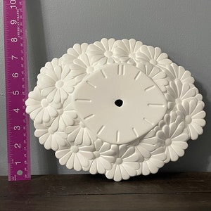 May include: White ceramic clock face with a raised daisy design. The clock face is blank and ready to be painted.