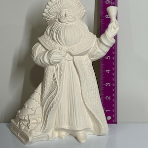 May include: An unpainted ceramic Santa Claus figure, featuring a long white beard and hat, holding a small bell. The figure is approximately 23 cm tall. The design includes detailed textures.