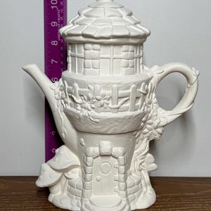 Könnte beinhalten: Weiße Keramik-Teekanne in Form eines Feenhauses mit einem Pilzfuß. Die Teekanne hat einen Schornstein, Fenster und eine Tür.