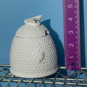 Op de afbeelding: Witte keramische honingpot met een honingraatpatroon en een bijenontwerp. Het deksel heeft een bijvormige handgreep. De honingpot is ongeveer 10 cm hoog.