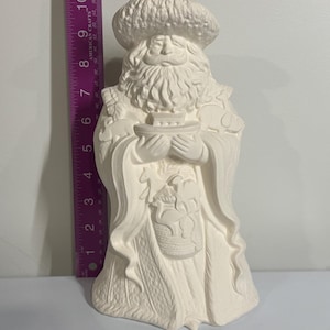 Könnte beinhalten: Eine weiße Keramikfigur des Weihnachtsmanns, etwa 25 cm hoch, die eine kleine Schale hält. Die Figur hat einen langen weißen Bart, einen Hut und einen Umhang. Die Details sind in die Keramik geschnitzt.