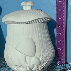 Puede incluir: Un recipiente blanco de cerámica con forma de hongo y tapa. El recipiente tiene una superficie texturizada y presenta diseños de hongos en relieve. Mide aproximadamente 20 cm de alto.