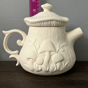 Puede incluir: Tetera de cerámica blanca con un diseño de setas. La tetera tiene una tapa y un asa. Las setas están en relieve en la tetera.