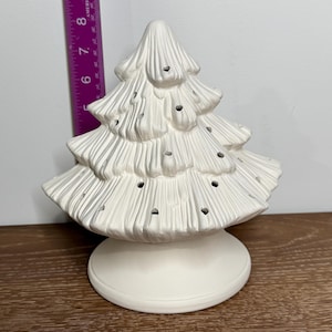 Op de afbeelding: Een witte keramische kerstboom met een ronde basis. De boom heeft meerdere niveaus met kleine gaten voor lichtjes. De boom is ongeveer 20 cm hoog.