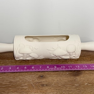 May include: A cream-colored ceramic rolling pin with a rectangular opening and mushroom designs. The rolling pin is approximately 15 inches long, with a purple ruler placed below it for scale.