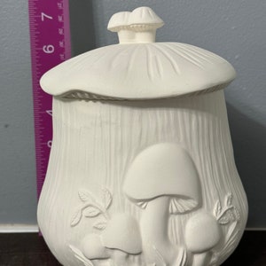 May include: A white ceramic mushroom-shaped container with a lid. The container has a textured surface and is decorated with embossed mushrooms and leaves. The lid is topped with two small mushroom shapes. The container is approximately 7 inches tall.